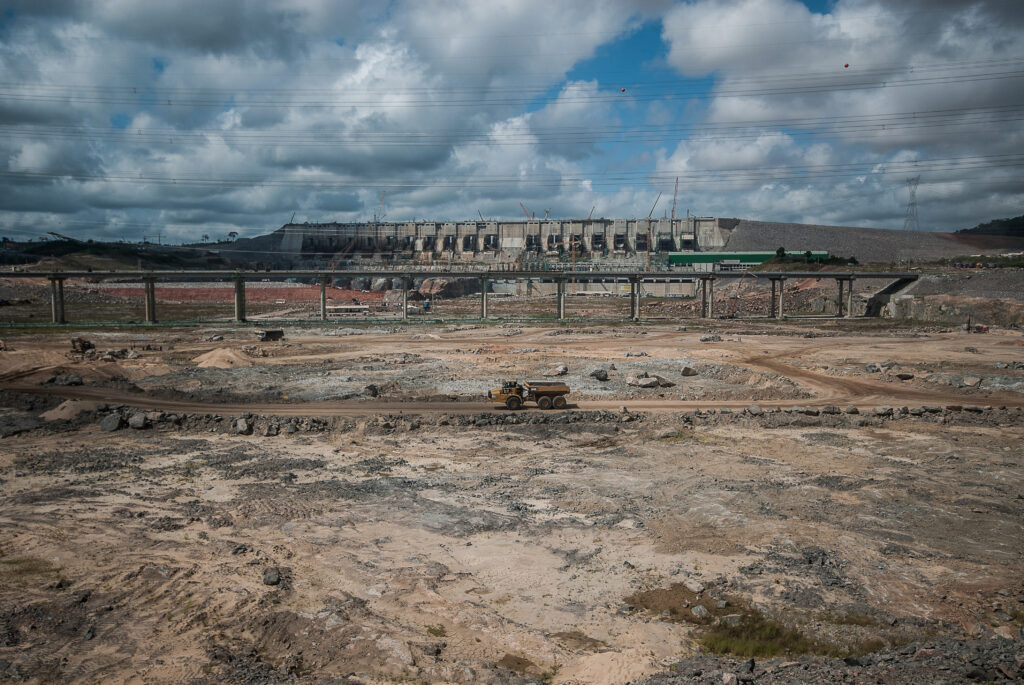 Canteiro de construção de Belo Monte. Terra avermelhada na frente. Ao fundo, a estrutura de concreto da hidrelétrica