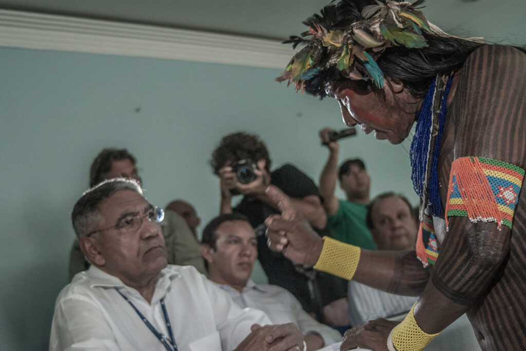 Um indígena com dedo em riste e em pé fala com um homem de camisa social sentado