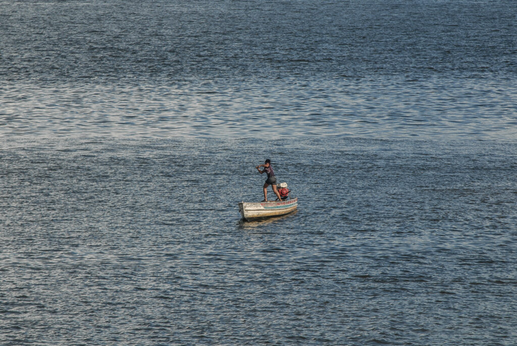 Um rio ocupa toda a foto. No meio da imagem, há um um homem em cima de um barco, pequeno comparado ao rio.