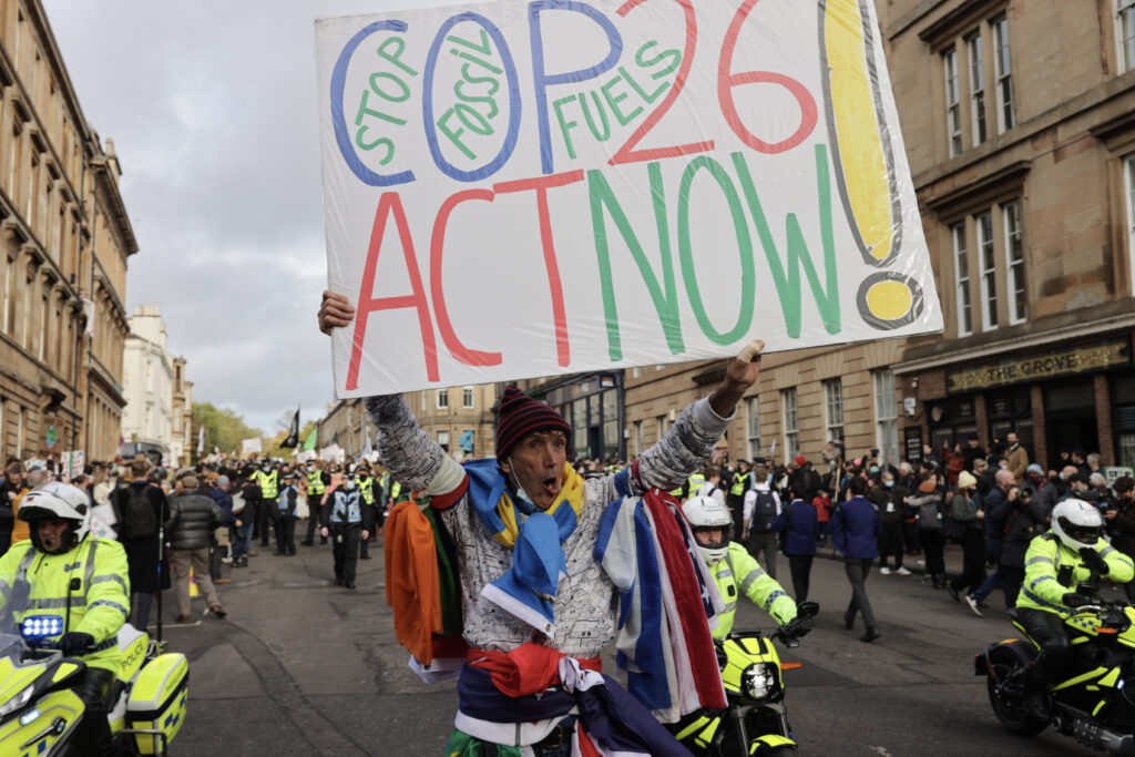 Um homem, envolto em bandeiras, segura no alto um cartaz em inglês COP26, act now