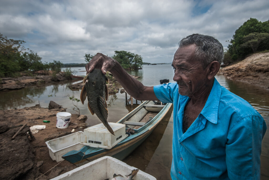 Um homem negro, em seus sessenta anos, segura um peixe. Ao fundo, um barco parado na beira do rio.
