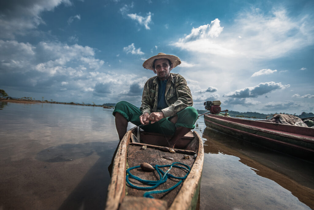 Um homem sentado em um pequeno barco. Ao fundo, o céu reflete na água do rio.