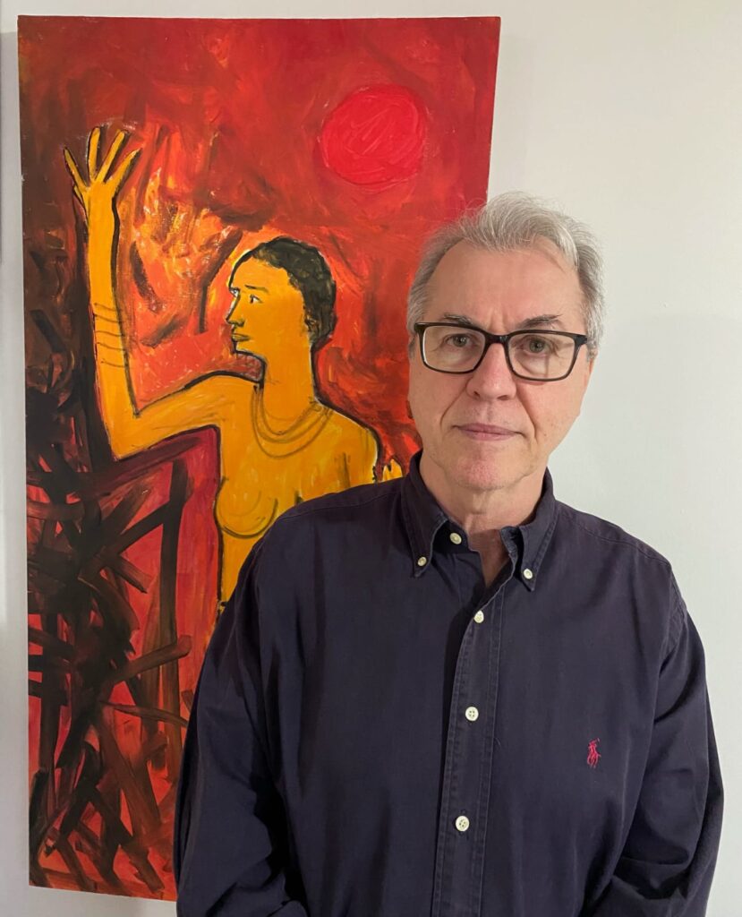 Ricardo Resende Figueira, em pé, ao lado de um quadro. Ele é branco, em seus 60 anos, usa óculos e tem cabelo branco