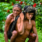 imagem de um indígena jovem carrega um idoloso nas suas costas. Ao fundo, há vegetação