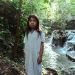 Uma criança indígena veste uma túnica branca em meio a uma floresta