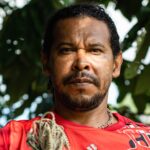 Um homem quilombola da Amazônia olha diretamente para a câmera, segurando uma rede de pescar. Ele tem pele, cabelos e olhos escuros, e veste uma camiseta vermelha. Ao fundo, uma copa de árvores.