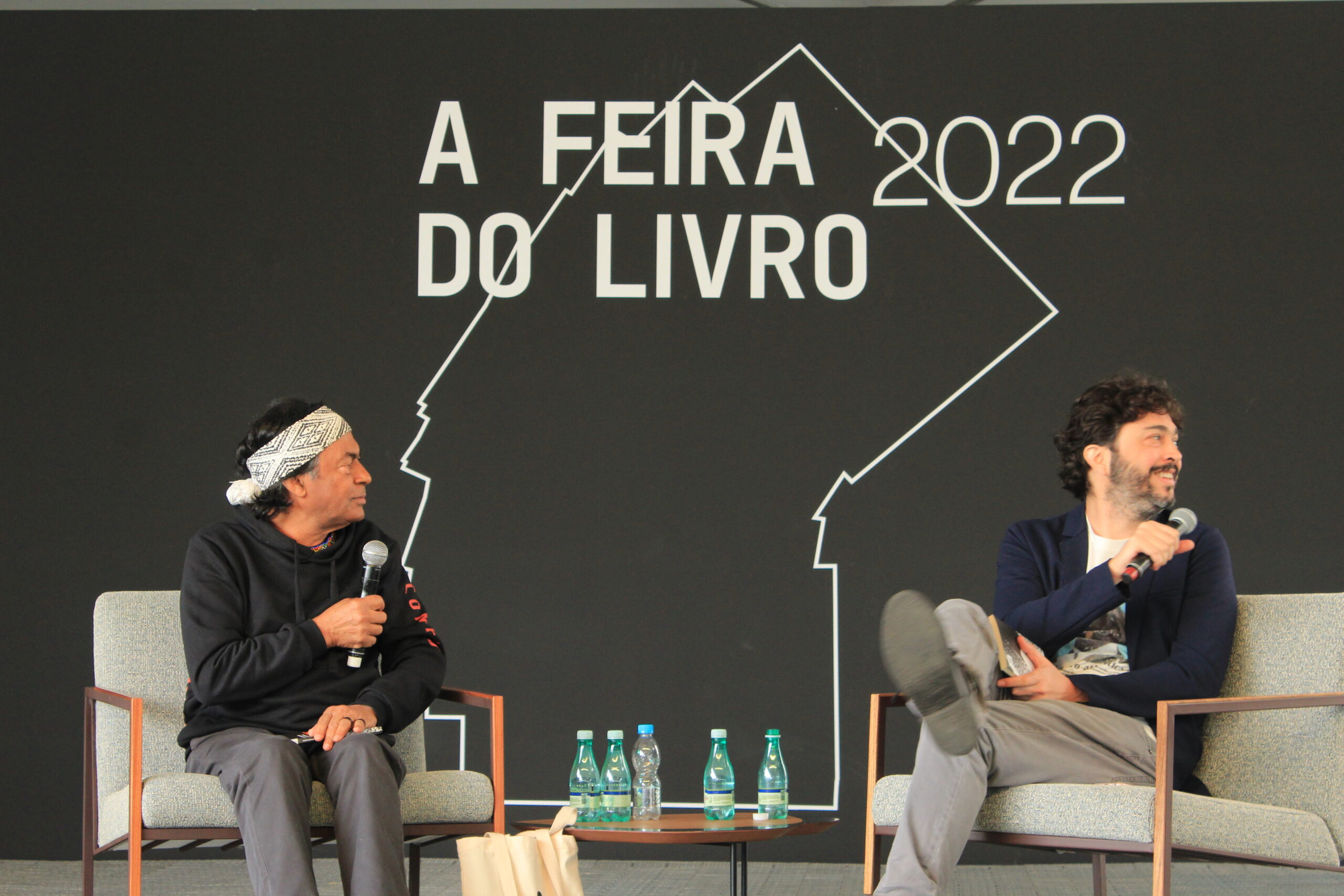 Ailton Krenak está à esquerda da foto, sentado, com um microfone na mão. Paulo Weneck está à direita, também sentado com um microfone, olhando sobre o ombro. Os dois discutem sobre patrimônio em um painel da Feira do Livro no Pacaembu, em São Paulo.