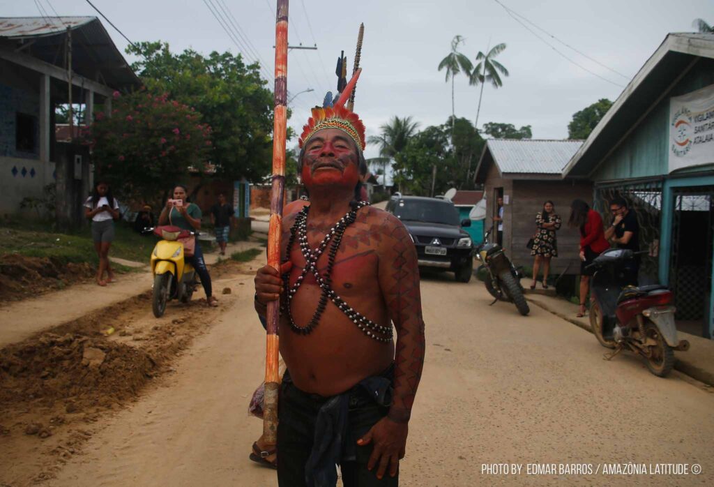Um indígena de cocar vermelho, segura um cajado colorido em uma rua de terra.